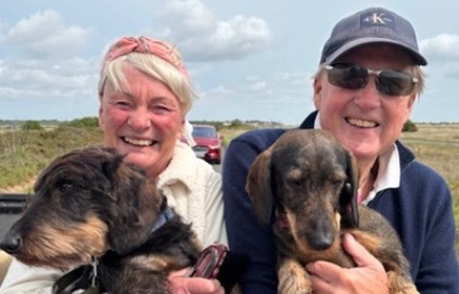 An older man and woman (Hugo and his wife) each holding a dog and sitting in the back of a car with an open roof. Hugo is wearing a baseball cap and sunglasses and his wife has a pink headband and blonde hair 
