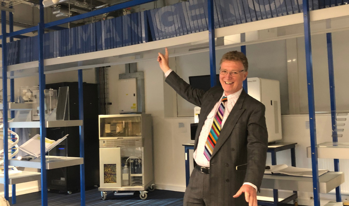 A man in a gray suit with a colorful striped tie smiles and points upward inside a laboratory-like setting. Behind him are various scientific instruments and machines.