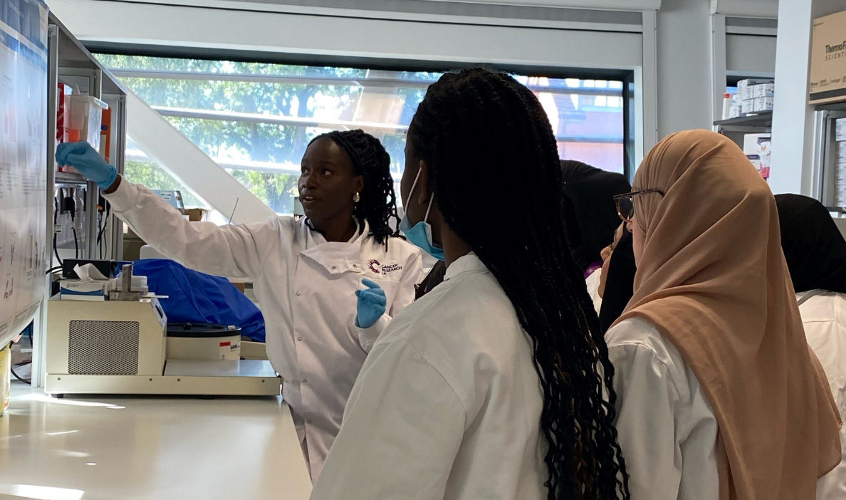 A woman in a white lab coat and blue gloves gestures toward a scientific poster while explaining it to a group of young women, also in lab coats, in a modern laboratory setting.