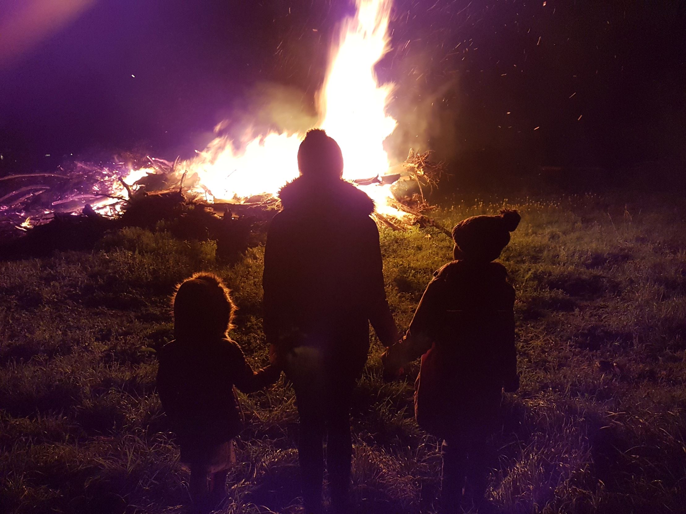 a woman standing looking at a bonfire with one of her children on both sides of her. She is holding their hands and they are all wearing winter coats and standing on grass