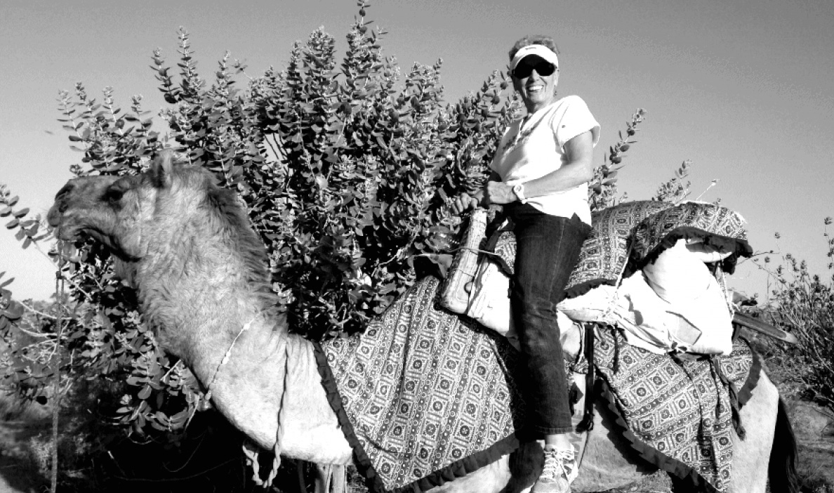 A woman wearing sunglasses, a visor, and casual clothes smiles while sitting on a decorated camel.