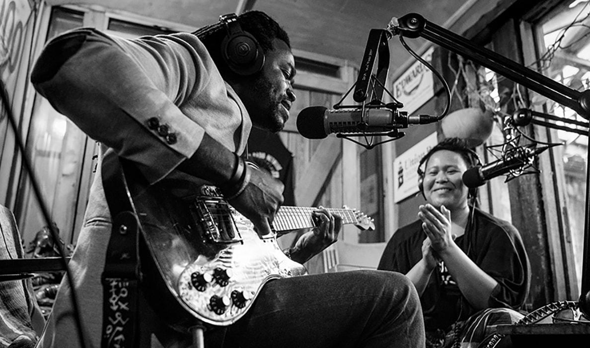 A man plays an electric guitar and sings into a microphone while wearing headphones in a cozy, intimate studio setting.