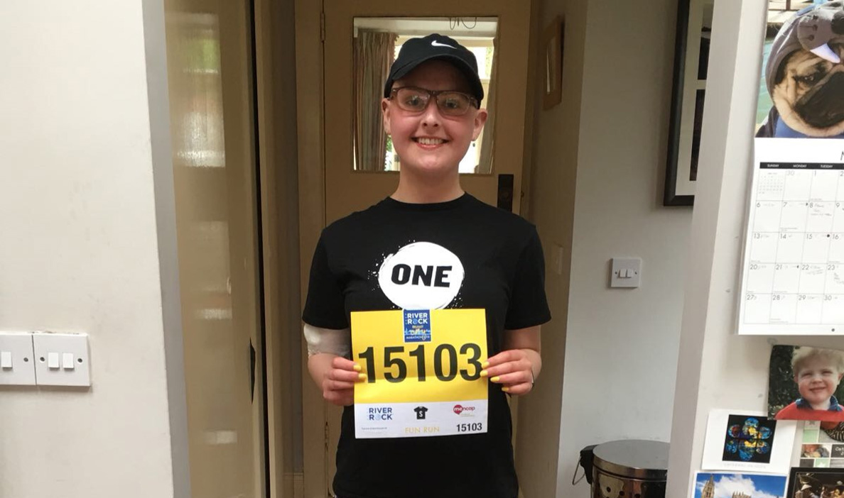 A young person stands indoors smiling and holding a yellow race bib with the number 15103. They are wearing a black cap, glasses, and a black t-shirt with the word "ONE" in bold letters.