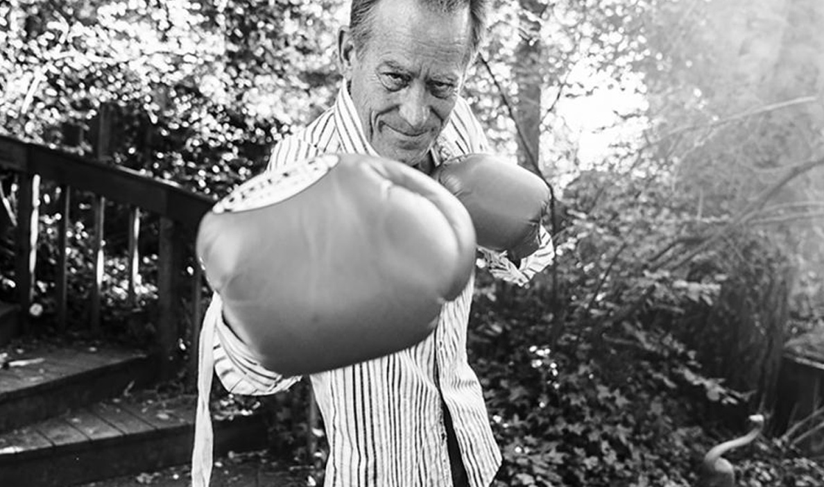 A man in a striped button-down shirt playfully poses with oversized boxing gloves in an outdoor setting surrounded by trees and foliage.