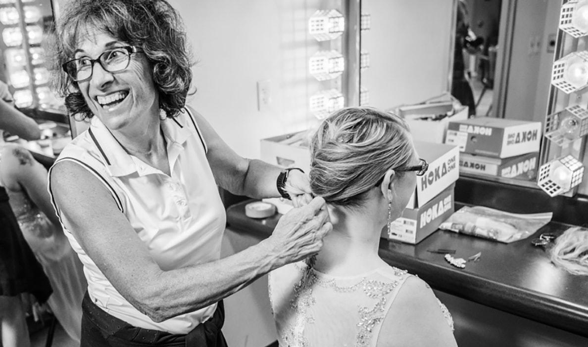 A woman smiles brightly as she styles another woman's hair in a backstage dressing room.