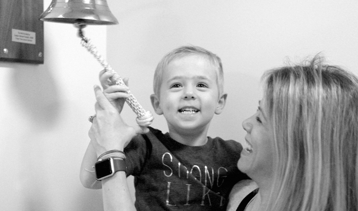 A smiling young boy rings a bell while being held up by his mother, who looks at him proudly.