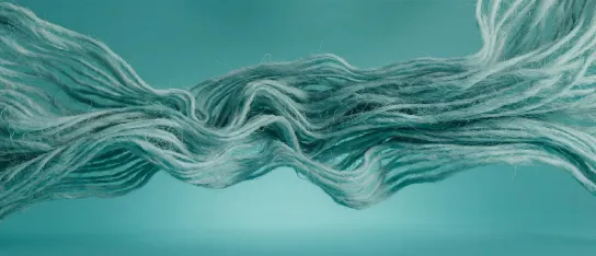 A landscape image depicting a bundle of flowing, fibrous strands resembling wool, thread, or natural fibres twisting and undulating horizontally across the frame. The strands are pale teal, white, and grey, set against a soft teal-blue background. 