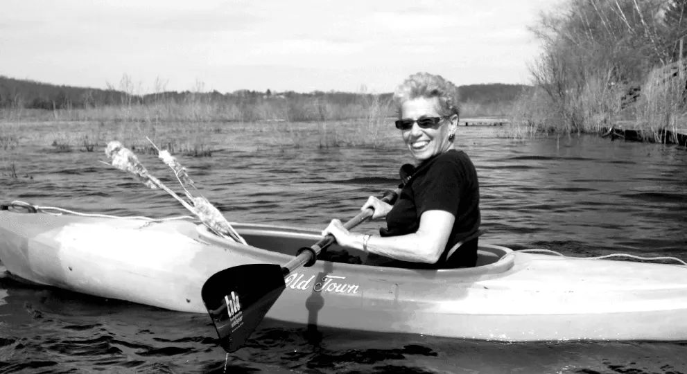 A woman wearing sunglasses and a dark t-shirt smiles while paddling a kayak.