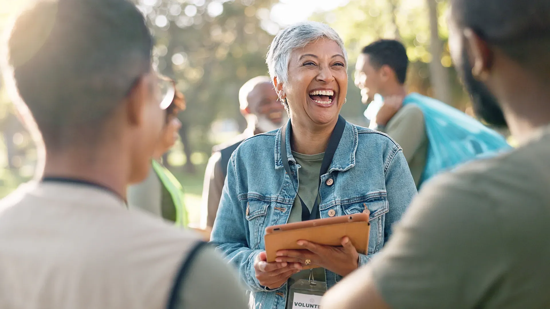 Smiling woman with short gray hair holding a tablet while talking to a support group outdoors.
