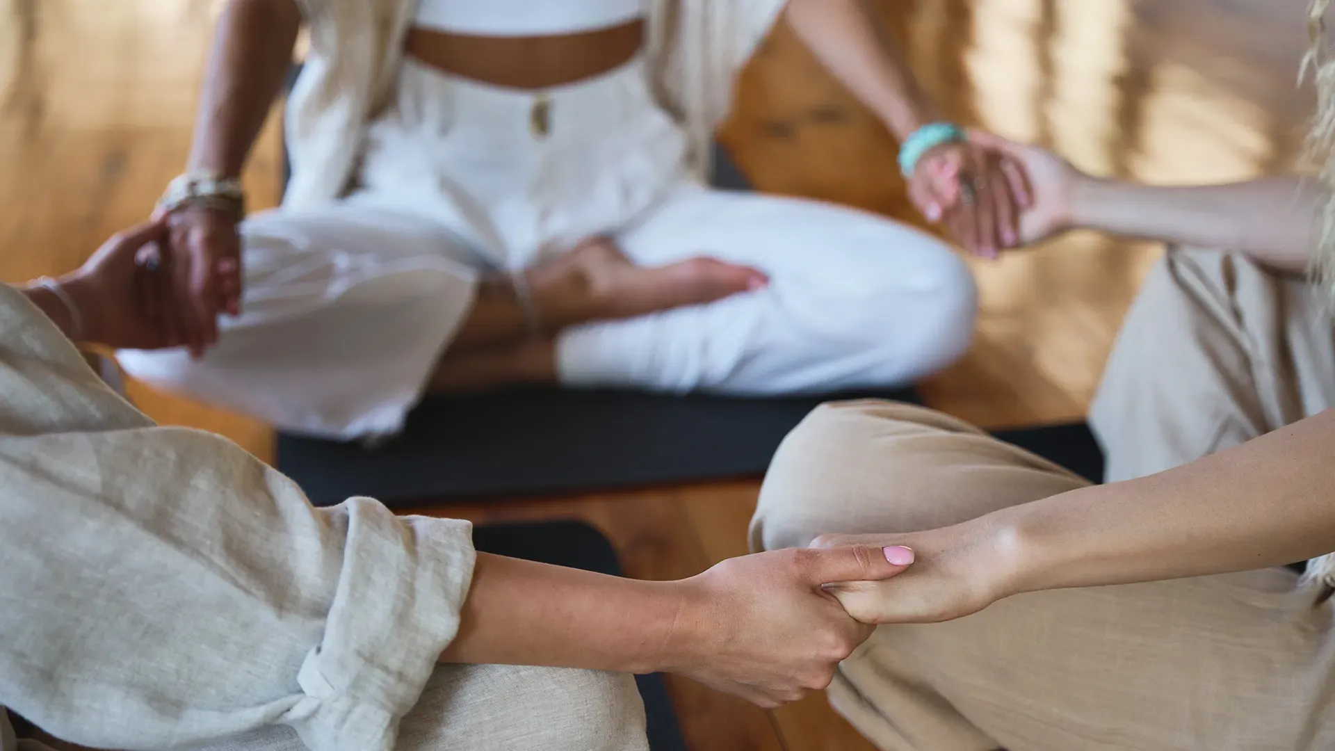 Group of people sitting cross-legged on yoga mats, holding hands in a supportive circle.