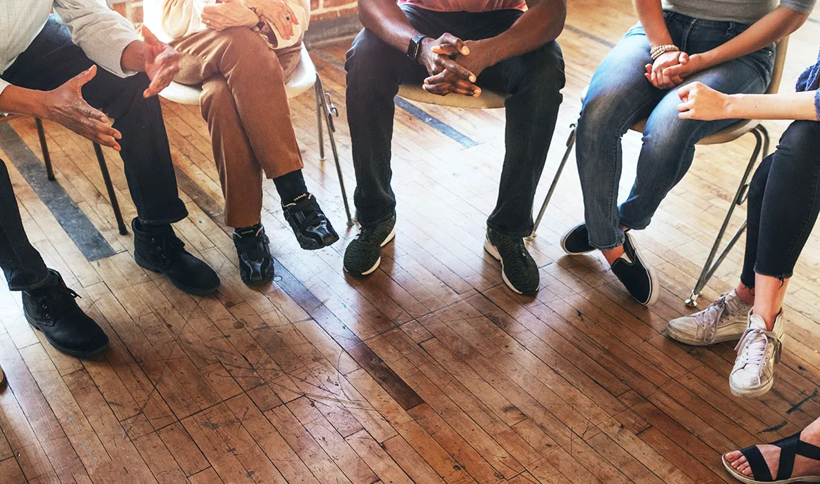 Diverse group of people sitting in a circle during a support or discussion group, only lower bodies visible.