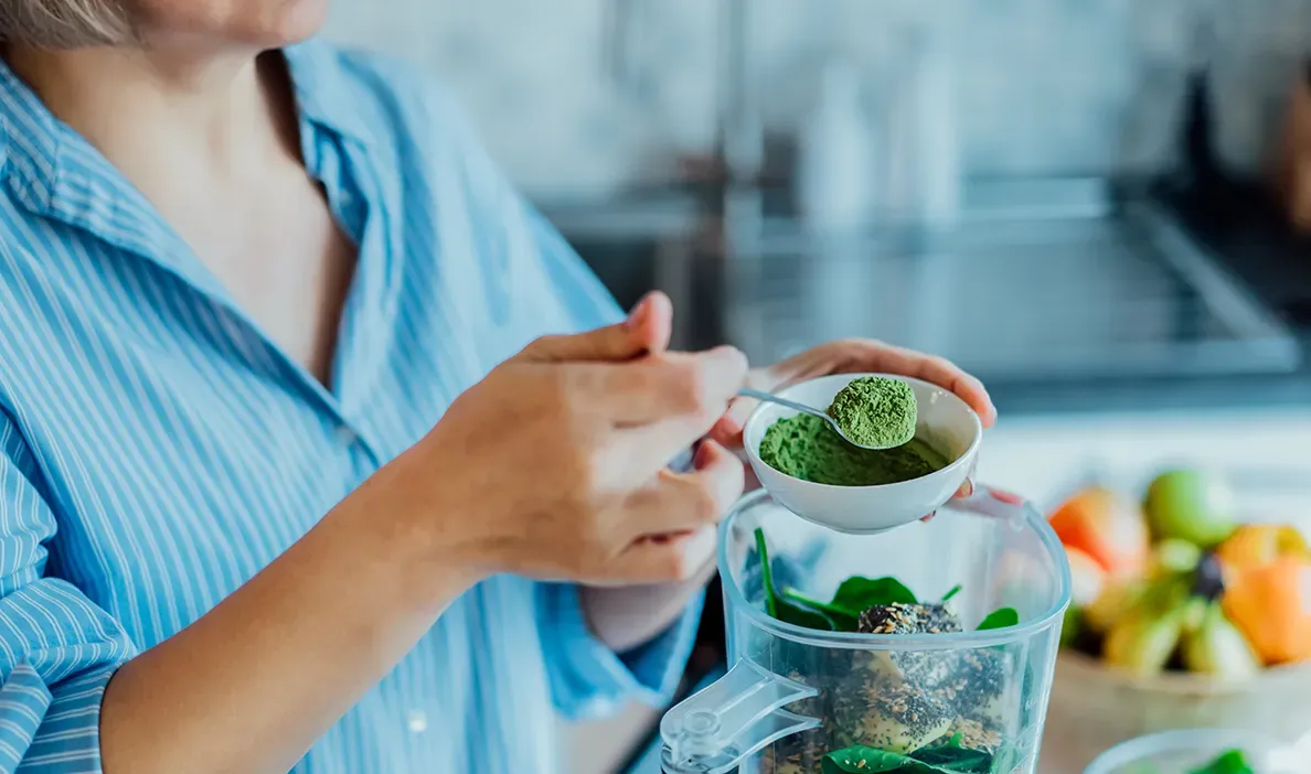 Woman preparing a healthy smoothie with green superfood powder in a blender.