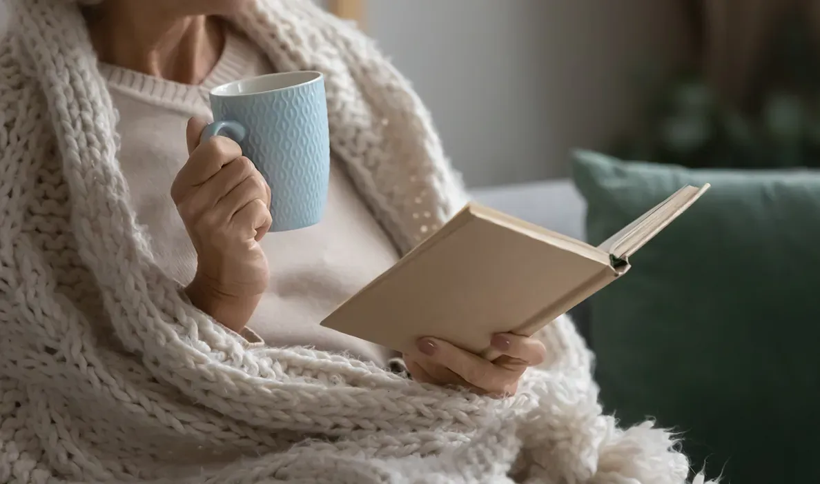 Woman wrapped in a cozy blanket, drinking from a blue mug while reading a book.