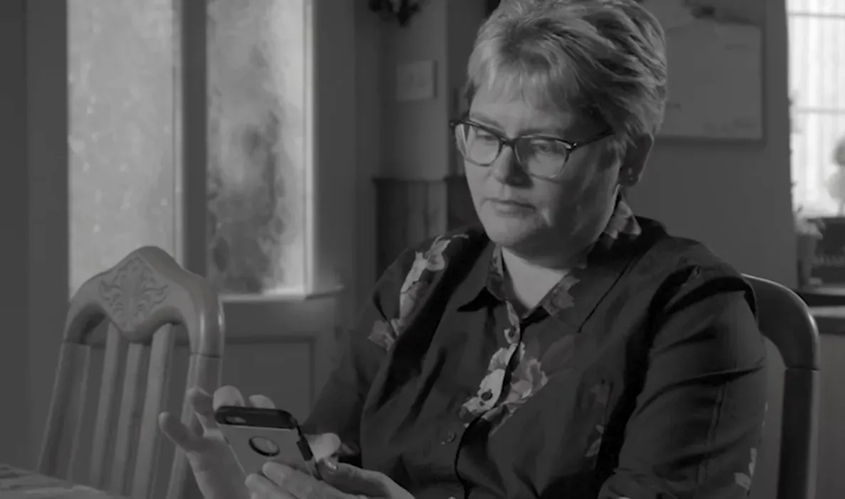 A woman with short, light-colored hair and glasses is sitting at a table, looking at a smartphone in her hands.