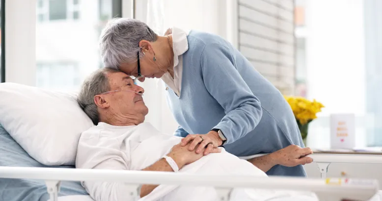 Person in a hospital bed holding hands with a visitor leaning close beside the bed.