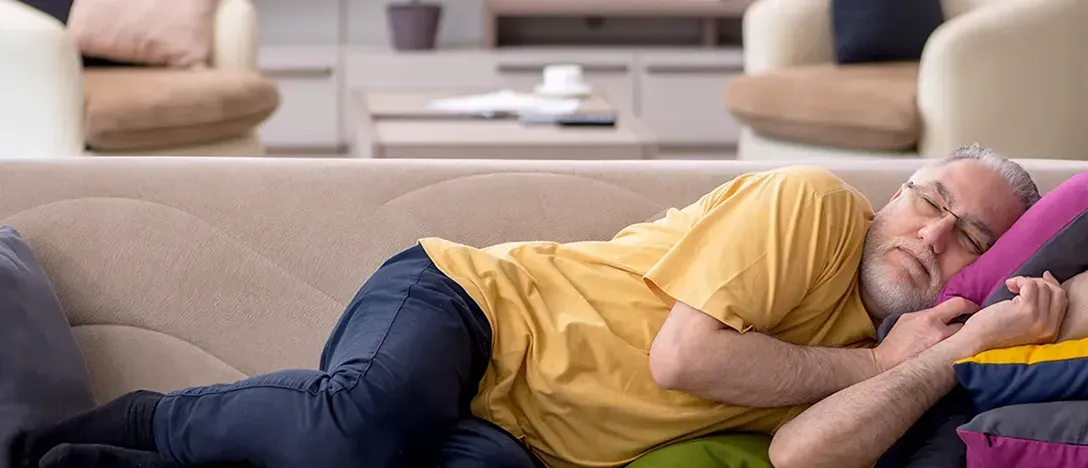 An elderly man with glasses and a beard rests on a couch, looking fatigued and weak.