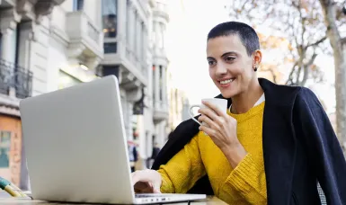 A woman with short hair and a bright smile sits at an outdoor café table, working on her laptop while holding a white coffee cup.