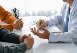 Doctor in a white coat discussing medical information with two patients at a table, with a clipboard, stethoscope, and paperwork visible.