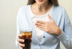 A woman in a light blue cardigan holds a glass of iced soda while placing a hand on her chest, indicating difficulty swallowing.