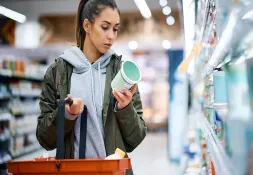 Woman shopping in a grocery store, examining a product label while holding a shopping basket.