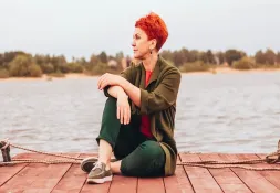 Woman with short red hair sitting on a wooden dock by a lake, gazing into the distance with a relaxed expression.
