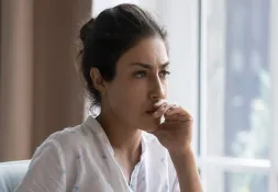 Woman with dark hair wearing a white shirt, sitting by a window with her hand on her mouth, appearing worried or pensive.
