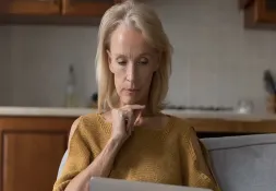 Woman with blonde hair wearing a mustard-colored sweater, sitting on a couch and thoughtfully looking at her laptop.