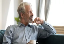 Man sitting on a couch, holding a white coffee mug and looking thoughtfully out the window.
