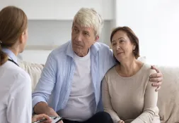 Senior couple sitting on a couch, smiling and listening attentively to a female doctor holding a clipboard during a consultation.
