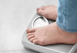 Person standing on a weighing scale, focusing on their feet and the scale&#039;s dial.
