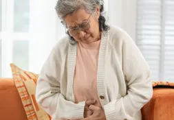Elderly woman sitting on a couch, clutching her stomach with a pained expression. 