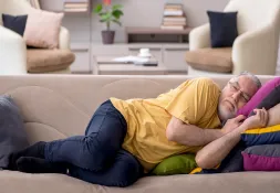 An elderly man with glasses and a beard rests on a couch, looking fatigued and weak.