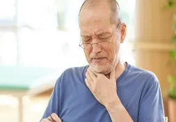 An elderly man wearing glasses and a blue shirt touches his throat with a pained expression.