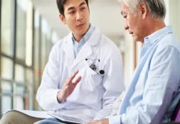 Doctor in a white coat talking to an older male patient, gesturing while discussing medical concerns.