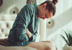 A young woman clutching her stomach in pain, sitting on a bed with a distressed expression.