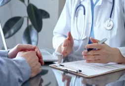Doctor in a white coat with a stethoscope discussing test results with a patient. The doctor&#039;s hands are gesturing while holding a pen.
