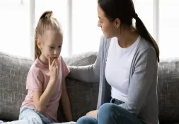 Mother sitting on a couch comforting her young daughter, who looks thoughtful and concerned.