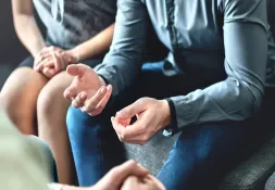Close-up of a man’s hands gesturing while discussing a serious topic, with a supportive person sitting nearby.