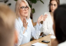 Professional woman with glasses and blonde hair speaking in a business meeting, gesturing with her hands.