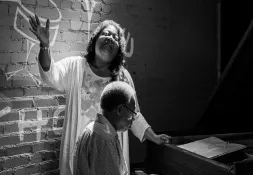Black and white photo of a woman singing expressively beside a man playing the piano, both bathed in stage lighting against a brick wall.