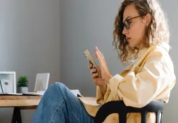 Young woman with curly hair and glasses sitting in a chair, focused on her smartphone.