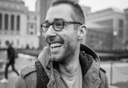 Black and white photo of a smiling man wearing glasses and a winter coat, outdoors with snowflakes on his shoulders.