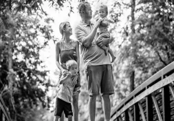  Black and white photo of a smiling family with two young children walking on a wooded footbridge, the father holding a baby and the mother beside their older child.