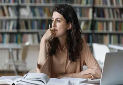 Woman sitting at a library table, looking thoughtful with a pencil in hand, surrounded by books and a laptop.