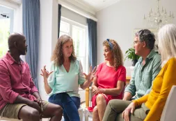 Middle-aged woman facilitating a group group session in a well-lit room with four attentive participants.