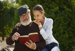 Elderly man reading a book while a young woman leans on his shoulder affectionately, both smiling outdoors.
