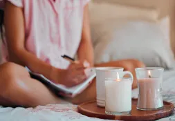 Person sitting cross-legged on a bed writing in a journal, with candles and a cup of tea on a wooden tray.