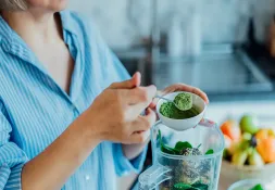 Woman preparing a healthy smoothie with green superfood powder in a blender.