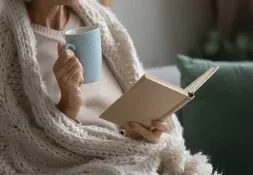 Woman wrapped in a cozy blanket, drinking from a blue mug while reading a book.