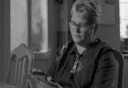 A woman with short, light-colored hair and glasses is sitting at a table, looking at a smartphone in her hands.